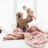 A mother holds her daughter while sitting with a Luxury Extra Large Oversized Throw Ballet Slipper (light pink) Colored Lush Saranoni Blanket. The soft blanket is an extra large throw blanket and has a soft faux fur feeling.