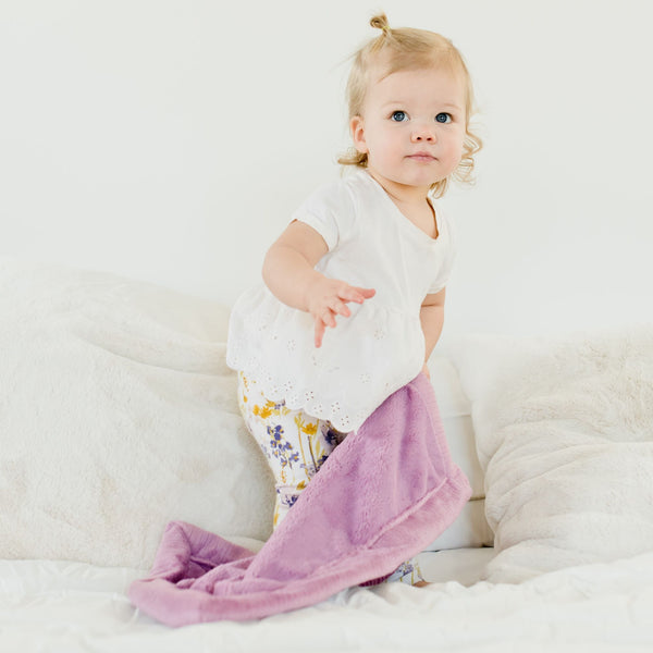 A little girl wearing a white outfit with floral pants holds a soft baby blanket that is a mini lush Saranoni Blanket. The Saranoni Blanket is a light purple with the specific color being called Fairy Wings.