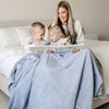 A mother sits with her son and daughter while holding a Luxury Extra Large Oversized Throw Storm Cloud (dusty blue gray) Colored Lush Saranoni Blanket. The soft blanket is an extra large throw blanket and has a soft faux fur feeling.