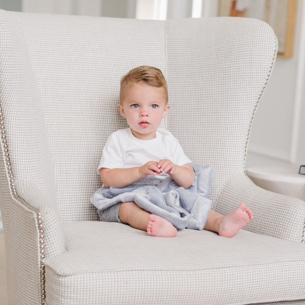 A little boy who is toddler aged with a white outfit on has a small soft light blue Saranoni Blanket draped over his lap. The soft blanket is a small blanket with lush fabric and the specific color is called Storm Cloud.