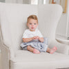 A little boy who is toddler aged with a white outfit on has a small soft light blue Saranoni Blanket draped over his lap. The soft blanket is a small blanket with lush fabric and the specific color is called Storm Cloud.