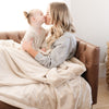 A woman holds her daughter while holding a Luxury Extra Large Oversized Throw Buttermilk (light tan) Colored Lush Saranoni Blanket. The soft blanket is an extra large throw blanket and has a soft faux fur feeling.