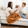 A mother holds her baby and has a Luxury Receiving Camel (light yellowish brown) Colored Lush Saranoni Blanket draped over her lap. The soft blanket is a small blanket and a baby blanket or toddler blanket.