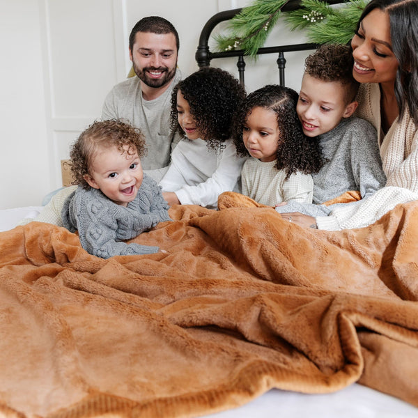 A husband and wife sit with their three daughters and son while holding a Luxury Extra Large Oversized Throw Camel (light yellowish brown) Colored Lush Saranoni Blanket. The soft blanket is an extra large throw blanket and has a soft faux fur feeling.