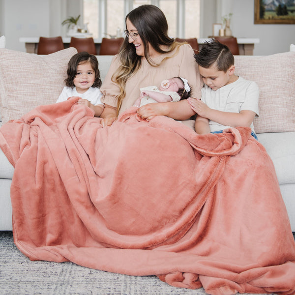 A mother sits with her son and two daughters while holding a Luxury Extra Large Oversized Throw Clay Colored Lush Saranoni Blanket. The soft blanket is an extra large throw blanket and has a soft faux fur feeling.