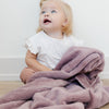 A little girl sits under a Luxury Receiving Bloom (dusty purple) Colored Lush Saranoni Blanket. The soft blanket is a small blanket and a baby blanket or toddler blanket.