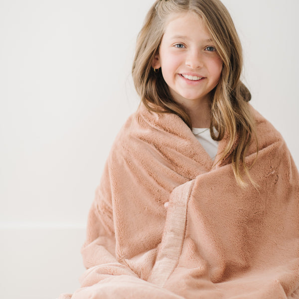 A little girl is wrapped in a Toddler Rosy (dusty peach) Colored Lush Luxury Saranoni Blanket. The soft blanket is a small blanket and a baby blanket or toddler blanket.