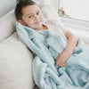 A little boy lays under a Luxury Toddler Dew (light blue) Colored Lush Saranoni Blanket. The soft blanket is a toddler blanket or baby blanket.