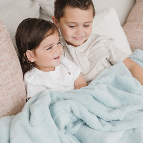 A brother and sister sit while holding a Luxury Extra Large Oversized Throw Dew (light blue) Colored Lush Saranoni Blanket. The soft blanket is an extra large throw blanket and has a soft faux fur feeling.