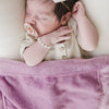 A baby girl in a tan outfit sleeps with a light purple soft Saranoni Baby Blanket draped over her. The blanket is a mini lush Saranoni Blanket and the specific color is called Fairy Wings.