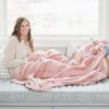 A woman sits on a window seat and holds a Ballet Slipper (light pink) Lush XL Saranoni blanket, designed for oversized comfort and featuring a buttery soft, plush texture. This luxurious Saranoni blanket is perfect for cozy evenings, reading, or home decor. The Saranoni blanket adds warmth, style, and unmatched softness, making it an ideal gift or essential for ultimate relaxation.