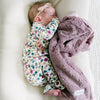 A baby girl lays by a Bloom (dusty purple) Colored Lush Saranoni Blanket. The soft blanket is a small blanket and a baby blanket or toddler blanket.