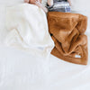 Two babies lay under two blankets. One is Camel Colored and one is an off-white. The specific color is called Natural. The Lush Saranoni Blankets are a soft blanket and a small blanket.