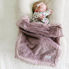 A baby girl lays under a Bloom (dusty purple) Colored Lush Saranoni Blanket. The soft blanket is a small blanket and a baby blanket or toddler blanket.