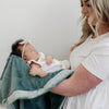 A mother holds her little girl in a Luxury Receiving Eucalyptus Colored Lush Saranoni Blanket. The soft blanket is a small blanket and a baby blanket or toddler blanket.