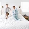 A little boy wrapped in a Luxury Receiving Dew (light blue) Colored Lush Saranoni Blanket plays on a bed with his brother. The soft blanket is a small blanket and a baby blanket or toddler blanket.