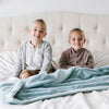 Two brothers sit while holding a Luxury Extra Large Oversized Throw Dew (light blue) Colored Lush Saranoni Blanket. The soft blanket is an extra large throw blanket and has a soft faux fur feeling.