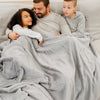 A father and his son and daughter sit while holding a Luxury Extra Large Oversized Throw Gray Colored Lush Saranoni Blanket. The soft blanket is an extra large throw blanket and has a soft faux fur feeling.