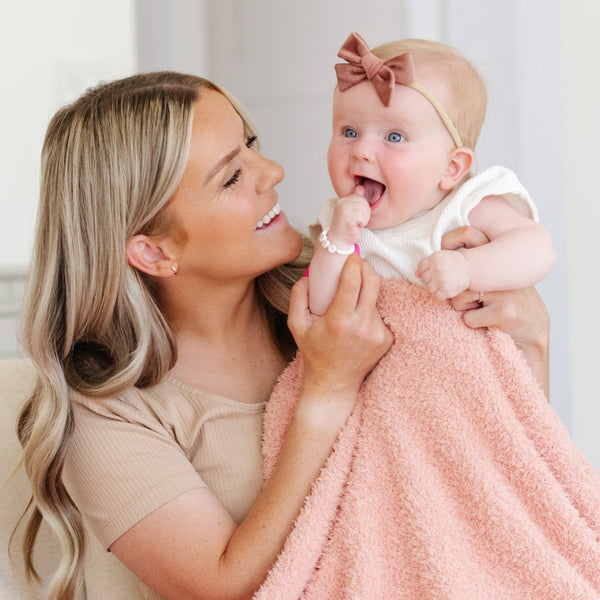 A mother holds her little girl and a peach blanket that is a Bamboni® blanket, offering ultimate softness and warmth. Made from Saranoni’s signature stretchy, cloud-like Bamboni® fabric, this luxury baby blanket provides comfort for naps, tummy time, and on-the-go snuggles. The soft blanket is a mini blanket and Saranoni blanket. 