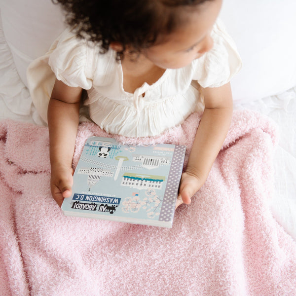 Little girl reading book on bed, snuggled in Light Pink BAMBONI TODDLER BLANKETS - Saranoni