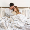 Husband and wife snuggling together in bed with a cozy blue striped bamboo muslin quilt. 