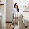 A woman with long dark hair in a white top and jeans stands in a light-colored kitchen holding a cream and tan striped blanket - Saranoni