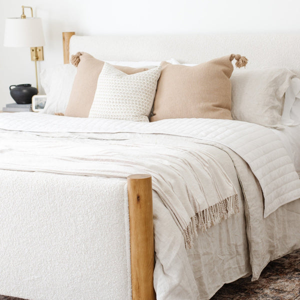 Close-up of a cream-colored bed with white sheets and pillows, accented by beige throw pillows and a striped blanket at the foot- Saranoni