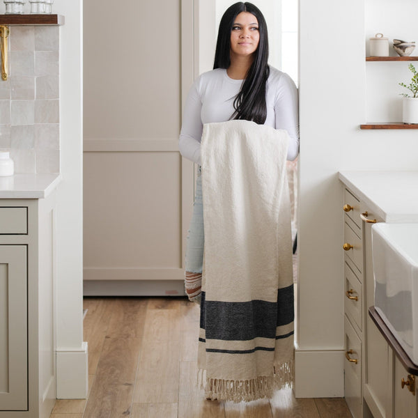 Woman with long dark hair in a light top and jeans stands in a bright kitchen holding a folded cream and gray striped blanket- Saranoni