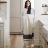 Woman with long dark hair in a light top and jeans stands in a bright kitchen holding a folded cream and gray striped blanket- Saranoni