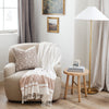 Decorative beige armchair with white throw blanket and stripes with a floral pillow next to wooden side table - Saranoni