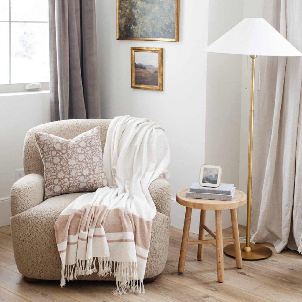 Decorative beige armchair with white throw blanket and stripes with a floral pillow next to wooden side table - Saranoni