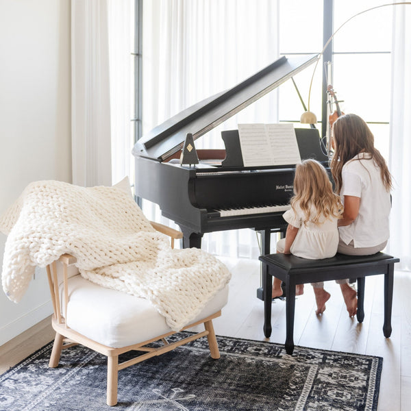 A mother and daughter play the piano by a soft, white Saranoni chunky knit throw blanket. The large knit Saranoni blanket, is made from 100% poly microfiber and drapes beautifully. This luxury throw blanket is both trendy and durable, making it the perfect addition to any space. Ideal for home decor enthusiasts and those seeking a cuddly, soft blanket, this chunky knit blanket combines style and comfort. Keywords: chunky knit throw, soft blanket, knit blanket, large knit blanket, luxury throw blanket.