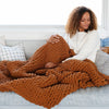 A woman sits under a soft, ginger colored Saranoni chunky knit throw blanket. The large knit Saranoni blanket, is made from 100% poly microfiber and drapes beautifully. This luxury throw blanket is both trendy and durable, making it the perfect addition to any space. Ideal for home decor enthusiasts and those seeking a cuddly, soft blanket, this chunky knit blanket combines style and comfort. Keywords: chunky knit throw, soft blanket, knit blanket, large knit blanket, luxury throw blanket.