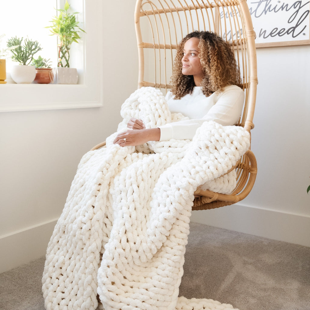 A woman relaxes in a wicker chair, wrapped in a soft, white Saranoni chunky knit throw blanket. The large knit Saranoni blanket, is made from 100% poly microfiber and drapes beautifully. This luxury throw blanket is both trendy and durable, making it the perfect addition to any space. Ideal for home decor enthusiasts and those seeking a cuddly, soft blanket, this chunky knit blanket combines style and comfort. Keywords: chunky knit throw, soft blanket, knit blanket, large knit blanket, luxury throw blanket.