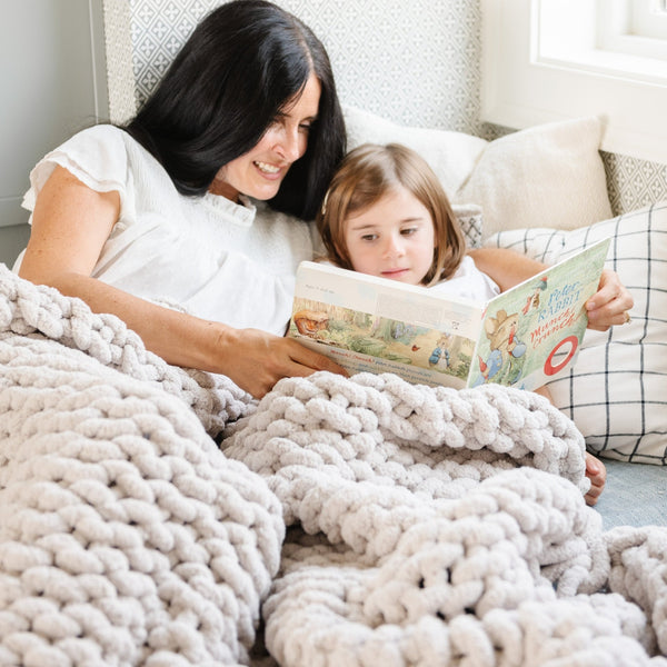 A mother and daughter sit under a soft, light gray colored Saranoni chunky knit throw blanket. The large knit Saranoni blanket, is made from 100% poly microfiber and drapes beautifully. This luxury throw blanket is both trendy and durable, making it the perfect addition to any space. Ideal for home decor enthusiasts and those seeking a cuddly, soft blanket, this chunky knit blanket combines style and comfort. Keywords: chunky knit throw, soft blanket, knit blanket, large knit blanket, luxury throw blanket.