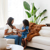 A mother and her children sit by a soft, ginger colored Saranoni chunky knit throw blanket. The large knit Saranoni blanket, is made from 100% poly microfiber and drapes beautifully. This luxury throw blanket is both trendy and durable, making it the perfect addition to any space. Ideal for home decor enthusiasts and those seeking a cuddly, soft blanket, this chunky knit blanket combines style and comfort. Keywords: chunky knit throw, soft blanket, knit blanket, large knit blanket, luxury throw blanket.