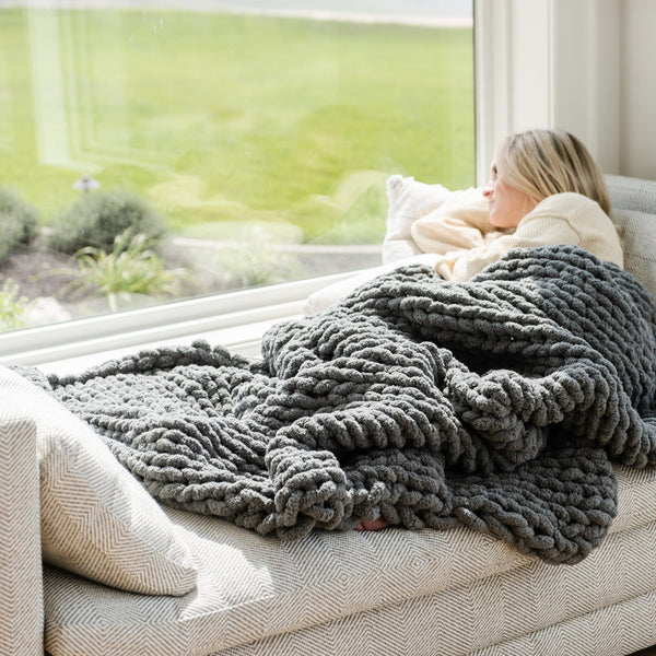 A woman lays under a soft, gray Saranoni chunky knit throw blanket. The large knit Saranoni blanket, is made from 100% poly microfiber and drapes beautifully. This luxury throw blanket is both trendy and durable, making it the perfect addition to any space. Ideal for home decor enthusiasts and those seeking a cuddly, soft blanket, this chunky knit blanket combines style and comfort. Keywords: chunky knit throw, soft blanket, knit blanket, large knit blanket, luxury throw blanket.