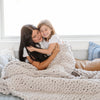 A mother and daughter sit under a soft, light gray colored Saranoni chunky knit throw blanket. The large knit Saranoni blanket, is made from 100% poly microfiber and drapes beautifully. This luxury throw blanket is both trendy and durable, making it the perfect addition to any space. Ideal for home decor enthusiasts and those seeking a cuddly, soft blanket, this chunky knit blanket combines style and comfort. Keywords: chunky knit throw, soft blanket, knit blanket, large knit blanket, luxury throw blanket.