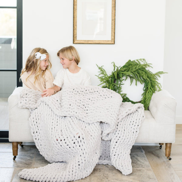 A sister and brother sit under a soft, light gray colored Saranoni chunky knit throw blanket. The large knit Saranoni blanket, is made from 100% poly microfiber and drapes beautifully. This luxury throw blanket is both trendy and durable, making it the perfect addition to any space. Ideal for home decor enthusiasts and those seeking a cuddly, soft blanket, this chunky knit blanket combines style and comfort. Keywords: chunky knit throw, soft blanket, knit blanket, large knit blanket, luxury throw blanket.