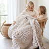 Two sisters sit under a soft, light gray colored Saranoni chunky knit throw blanket. The large knit Saranoni blanket, is made from 100% poly microfiber and drapes beautifully. This luxury throw blanket is both trendy and durable, making it the perfect addition to any space. Ideal for home decor enthusiasts and those seeking a cuddly, soft blanket, this chunky knit blanket combines style and comfort. Keywords: chunky knit throw, soft blanket, knit blanket, large knit blanket, luxury throw blanket.