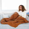 A woman sits under a soft, ginger colored Saranoni chunky knit throw blanket. The large knit Saranoni blanket, is made from 100% poly microfiber and drapes beautifully. This luxury throw blanket is both trendy and durable, making it the perfect addition to any space. Ideal for home decor enthusiasts and those seeking a cuddly, soft blanket, this chunky knit blanket combines style and comfort. Keywords: chunky knit throw, soft blanket, knit blanket, large knit blanket, luxury throw blanket.