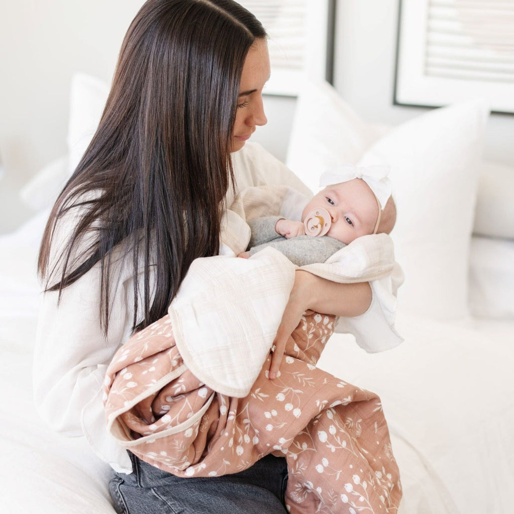 A mother holds her little girl in a Saranoni blanket. The baby is wrapped in a beautifully soft and breathable muslin quilt, designed for both comfort and style. The quilt features a warm, earthy rust tone with delicate white botanical accents, adding a timeless and elegant touch. Its airy, multi-layered muslin fabric is crafted to become even softer with each wash, making it a perfect choice for snuggles, naps, and everyday moments. The soft blanket is a luxury blanket and a Saranoni blanket. 