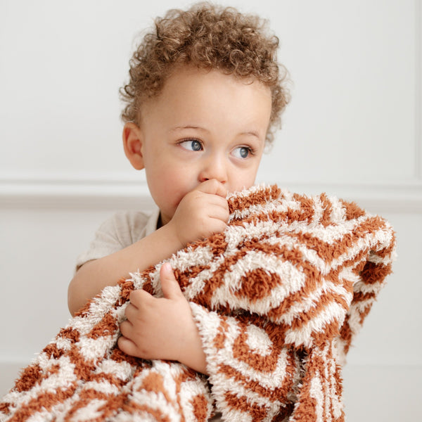 A little boy holds a double-layer Bamboni® baby blanket. The plush, breathable blanket featuring a chic burnt orange and cream geometric pattern, offering ultimate softness and warmth. Made from Saranoni’s signature stretchy, cloud-like Bamboni® fabric, this luxury baby blanket provides comfort for naps, tummy time, and on-the-go snuggles. The soft blanket is a kids blanket and a Saranoni blanket. 