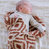 A baby boy lays in a double-layer Bamboni® baby blanket. The plush, breathable blanket featuring a chic burnt orange and cream geometric pattern, offering ultimate softness and warmth. Made from Saranoni’s signature stretchy, cloud-like Bamboni® fabric, this luxury baby blanket provides comfort for naps, tummy time, and on-the-go snuggles. The soft blanket is a mini blanket and Saranoni blanket. 