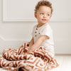 A little boy sits with a double-layer Bamboni® baby blanket. The plush, breathable blanket featuring a chic burnt orange and cream geometric pattern, offering ultimate softness and warmth. Made from Saranoni’s signature stretchy, cloud-like Bamboni® fabric, this luxury baby blanket provides comfort for naps, tummy time, and on-the-go snuggles. The soft blanket is a receiving blanket and Saranoni blanket. 