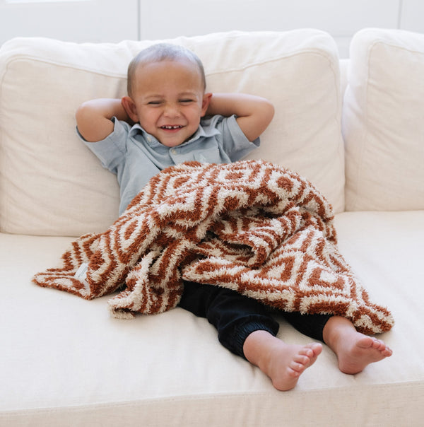 A little boy sits on a couch with a double-layer Bamboni® baby blanket. The plush, breathable blanket featuring a chic burnt orange and cream geometric pattern, offering ultimate softness and warmth. Made from Saranoni’s signature stretchy, cloud-like Bamboni® fabric, this luxury baby blanket provides comfort for naps, tummy time, and on-the-go snuggles. The soft blanket is a receiving blanket and Saranoni blanket. 