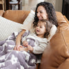 A mother sits with her little girl and a double-layer Bamboni® blanket. The plush, breathable blanket features a two-tone purple and white floral design, offering ultimate softness and warmth. Made from Saranoni’s signature stretchy, cloud-like Bamboni® fabric, this luxury baby blanket provides comfort for naps, tummy time, and on-the-go snuggles. The soft blanket is a receiving blanket and Saranoni blanket. 