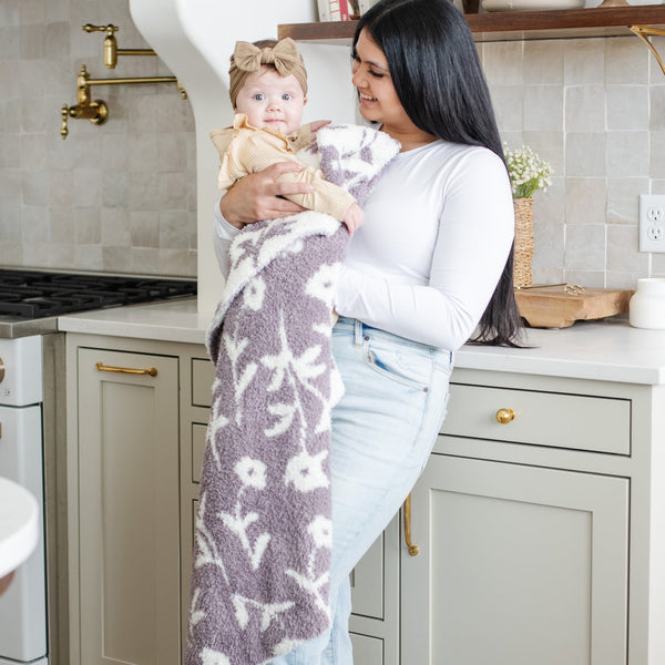 A mother smiles and holds her baby girl who is holding a double-layer Bamboni® baby blanket. The plush, breathable blanket features a two-tone purple and white floral design, offering ultimate softness and warmth. Made from Saranoni’s signature stretchy, cloud-like Bamboni® fabric, this luxury baby blanket provides comfort for naps, tummy time, and on-the-go snuggles. The soft blanket is a receiving blanket and Saranoni blanket. 