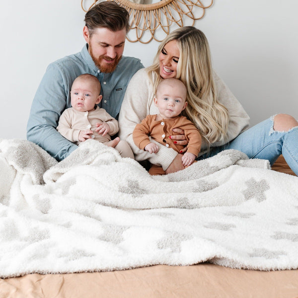 A husband and wife sit with their twin boys and a luxurious double-layer Bamboni® blanket by Saranoni, featuring a cream and taupe blanket with a swiss cross pattern. This ultra-soft blanket is crafted from bamboo rayon and poly microfiber, making it lightweight, breathable, and fluffy. Ideal as a cozy throw blanket, luxury blanket, plush throw, soft blanket, or stylish home accent, this Saranoni blanket brings warmth, comfort, and timeless elegance to any living room or bedroom.