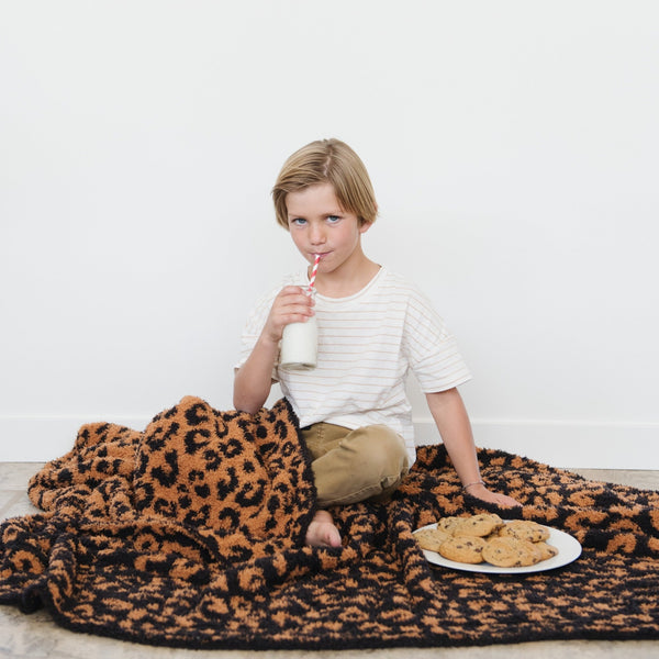 A little boy has cookies and milk and is with a luxurious double-layer Bamboni® blanket by Saranoni, featuring classic leopard print of orange and black. This ultra-soft blanket is crafted from bamboo rayon and poly microfiber, making it lightweight, breathable, and fluffy. Ideal as a cozy throw blanket, luxury blanket, plush throw, soft blanket, or stylish home accent, this Saranoni blanket brings warmth, comfort, and timeless elegance to any living room or bedroom.